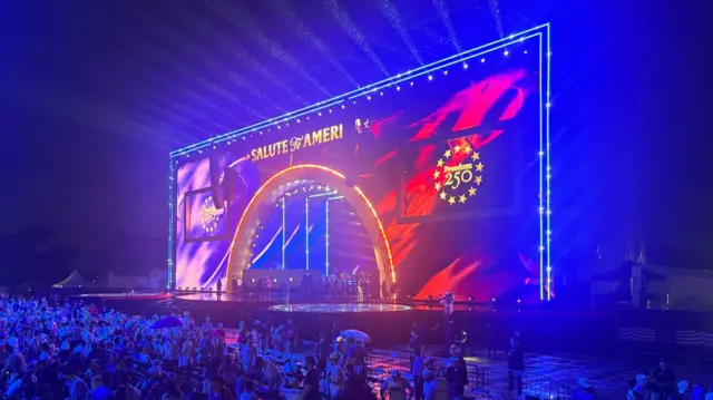 A large stage is lit up in blue, with signs reading "Salute to America" and "Freedom 250" visible.