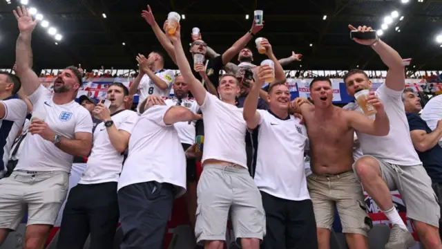 A group of England football fans are showing cheering with their arms around each other, holding drinks aloft.
