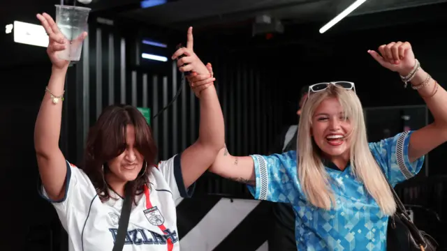 Two female fans in England football jerseys celebrating with their hands up in the air.