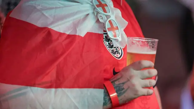A man holds a pint. He wears an England shirt and an England flag.