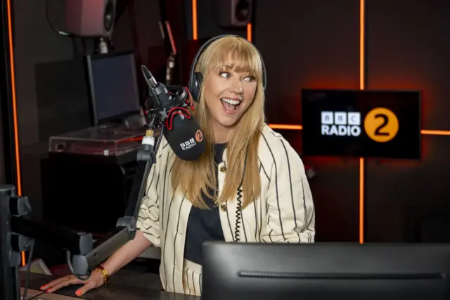 Sara Cox smiles as she sits in front of a BBC Radio 2 microphone in her studio