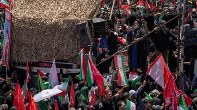 A member of the clergy stands on a platform and uses a microphone to talk to the crowds below