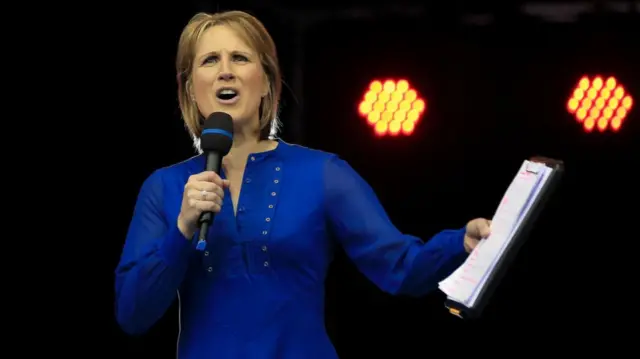 Katharine Merry is showing speaking with a microphone, with papers on a clipboard in her left hand.