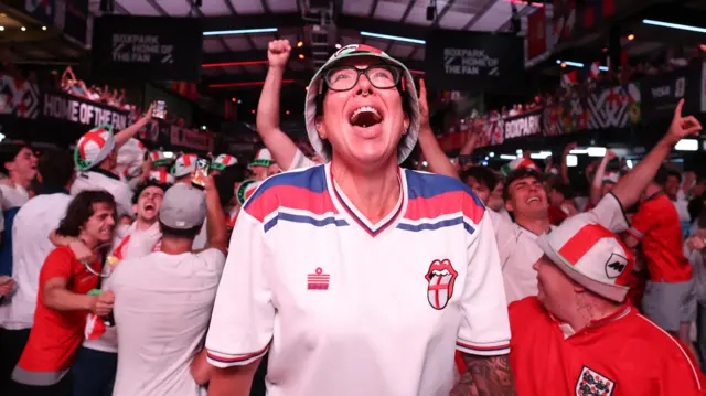 A fan celebrating wearing a bucket hat and England football jersey in a massive crowd of people.