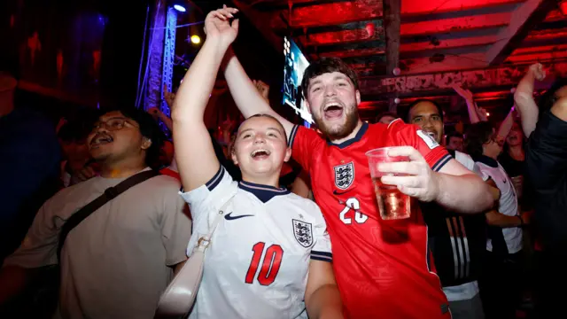 Two fans wearing England football jerseys celebrate with their arms in the air.