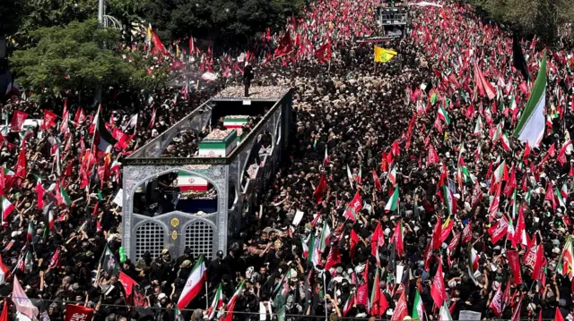 A vehicle carrying coffins makes its way through large scores of people