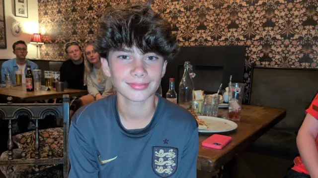 A teenage boy smiling into the camera wearing a blue England football jersey.