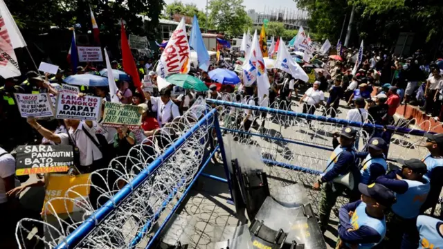 Protesters with colourful signs walk past a gate with barbed wire