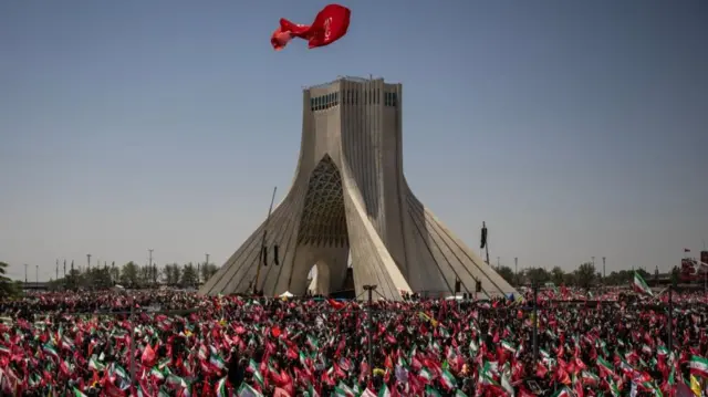 A flag flies above a monument with scores of people gathered below