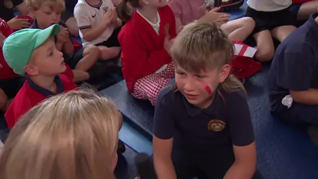 Student talking to BBC journalist in school uniform sitting on the floor with other students around