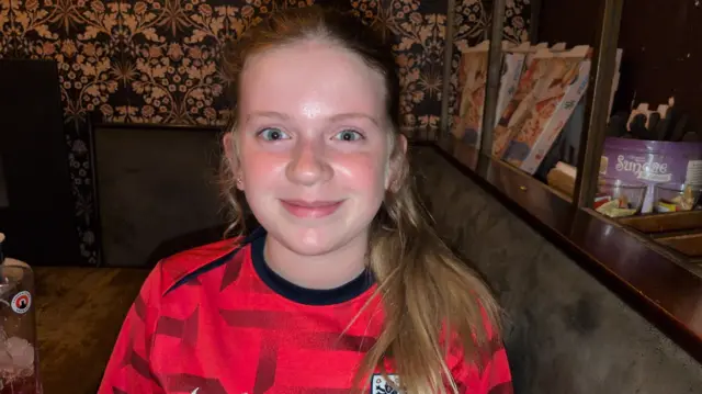 A teenage girl smiling into the camera wearing a red England football jersey.