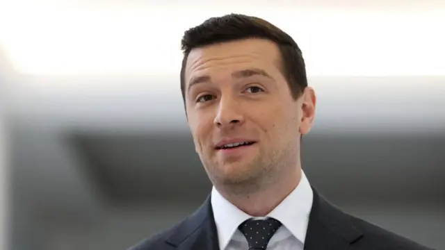 Jordan Bardella against a blurred background. He is wearing a dark coloured suit.