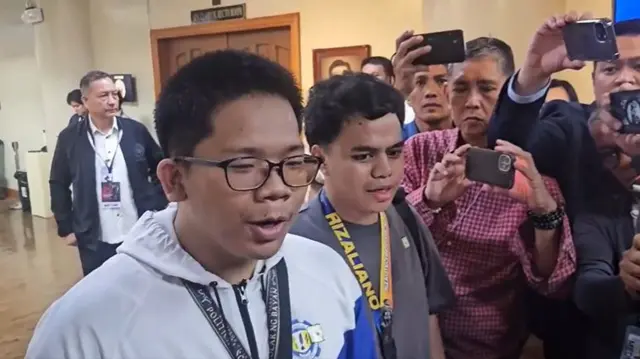 A young man with a white hoodie speaking to reporters