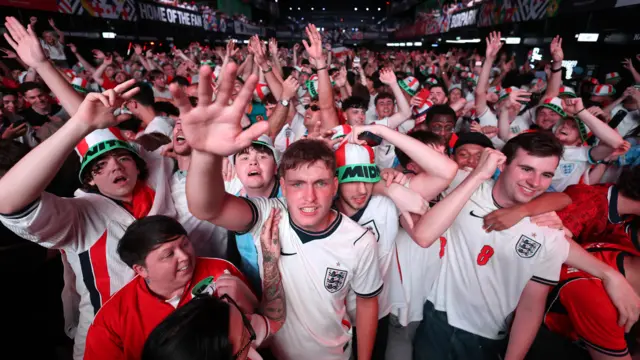 A group of male fans celebrate England's World Cup win with their hands raised in a crowd.