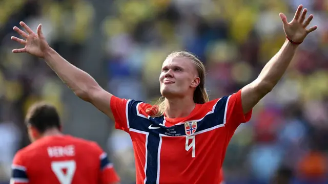 Erling Haaland in Norway's football kit with hands up celebrating on pitch.