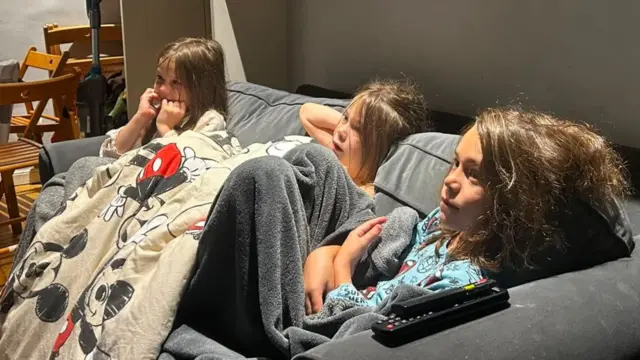Three kids watching the football under a blanket on a sofa.