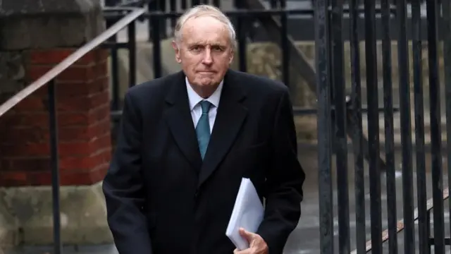 Paul Dacre walks through a black metal gate, carrying a piece of paper
