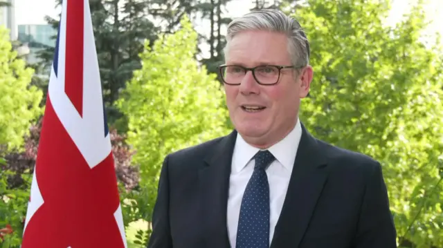 Keir Starmer speaks to broadcasters at the NATO summit in Turkey. He is standing next to a Union Jack flag with trees in the background