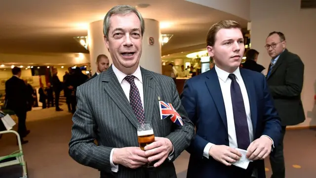Nigel Farage holds a beer at a European Parliament session in January 2020. He is standing next to George Cottrelll