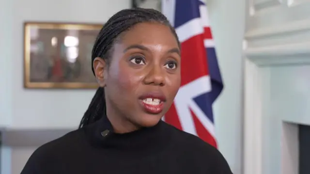 Kemi Badenoch is shown speaking, with a Union Jack flag behind her.