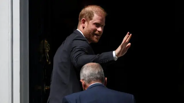 Prince Harry wears a suit and walks through a door, he waves at the camera