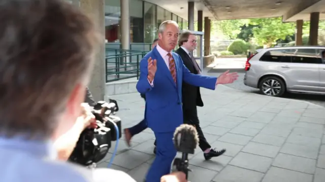 Nigel Farage walks in a deep blue suit. Members of the press appear to be gathered around him, holding out audio equipment.