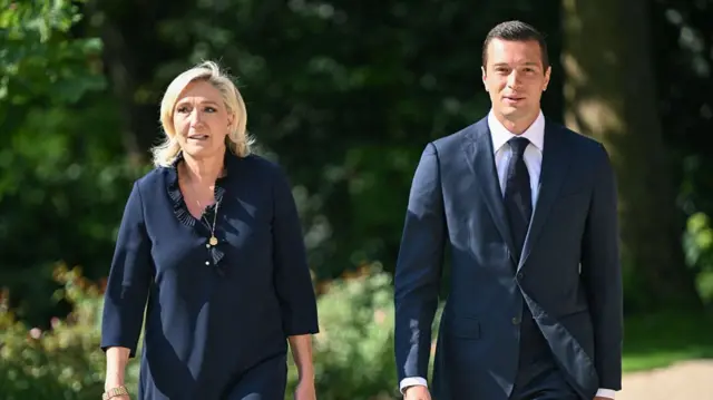 Marine Le Pen, a woman dressed in dark colours and wearing a necklace, on the left. Jordan Bardella, a man, wearing a black tie, white shirt and dark blazer, on the right. Behind the pair is greenery.