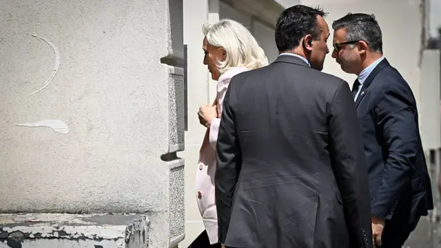 Marine Le Pen, a woman, entering a doorway. She is surrounded by men in suits,
