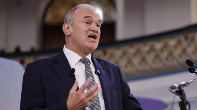 Ed Davey is pictured speaking behind a microphone, wearing a suit and tie.