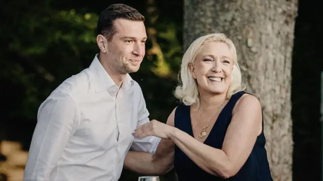 A young man in a white shirt stands on the left beside a smiling blonde-ahired woman in a black sleeveless dress