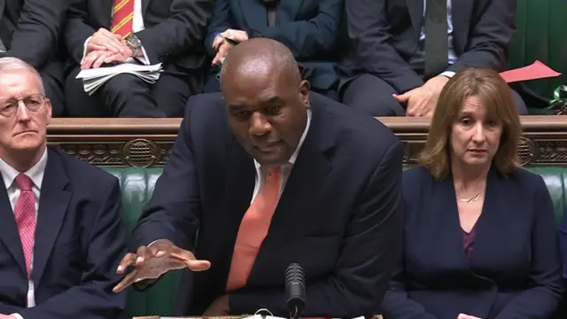 David Lammy in a dark suit answers questions in the Commons. Northern Ireland Secretary Hilary Benn (L) and Chancellor Rachel Reeves (R) sit on the leather green benches behind him