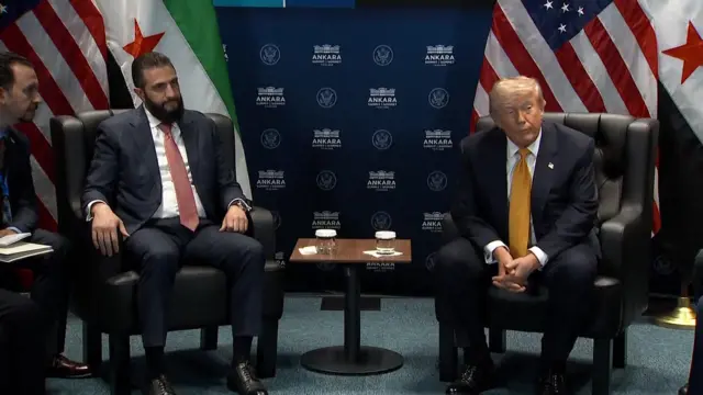 Donald Trump and Syrian President Ahmed al-Sharaa sit next to each other in large leather armchairs during a bilateral meeting at the Nato summit. Al-Sharaa has short black hair and a bushy beard and is wearing a navy suit with a white shirt and red tie. Trump is also wearing a navy suit and white shirt but with a yellow tie.