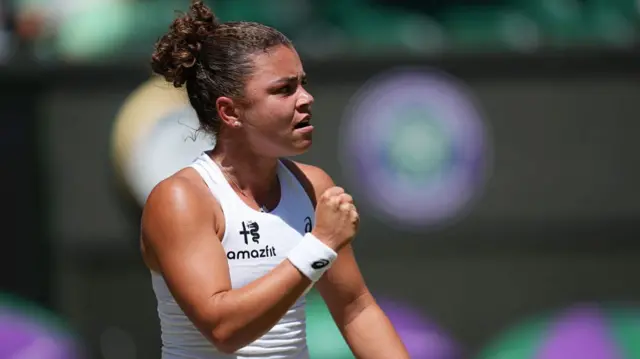 Jasmine Paolini, wearing a white sleeveless vest and her dark hair in a bun, raises a clenched right fist