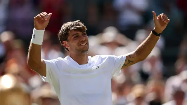 Flavio Cobolli, wearing a white t-shirt, raises his arms in the air with his thumbs up