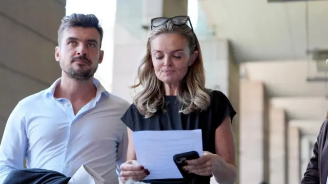 Laura Enston stands holding a sheet of paper while a man stands to her right