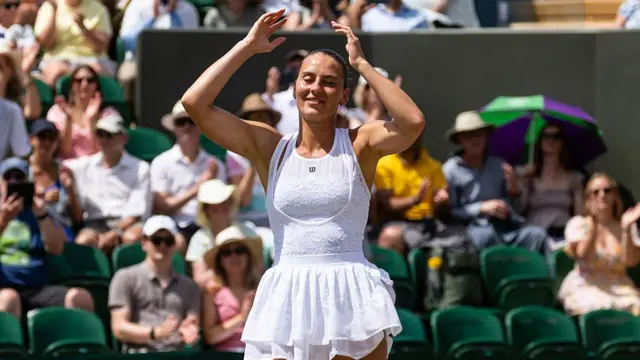 Marta Kostyuk, wearing a white sleeveless ballerina-style dress, raises her arms above her head in disbelief with the crowd blurred in the background