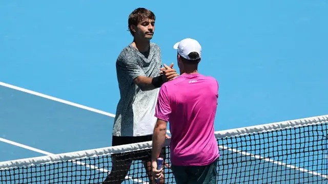 Flavio Cobolli, wearing a black and white patterned top, shakes hands with Arthur Fery, facing away from the camera with a white cap and purple t-shirt, at the net