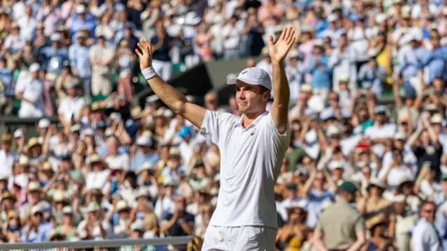 Arthur Fery, wearing a white polo shirt and cap, raises his arms in the air in front of a packed Centre Court crowd