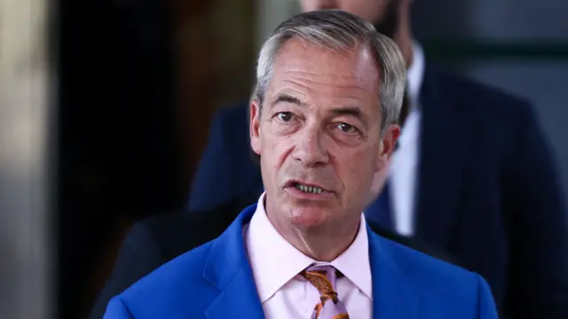 Close up of Nigel Farage in a royal blue suit, pink shirt and patterned tie (in pink and orange) mid-speech. A blurred figure in a black suit stands behind him in the background
