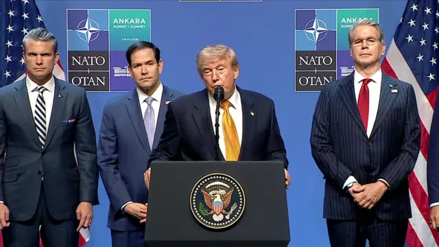 Donald Trump stands at a microphone wearing a dark suit and gold coloured tie. Officials including Pete Hegseth and Marco Rubio can be seen behind him.