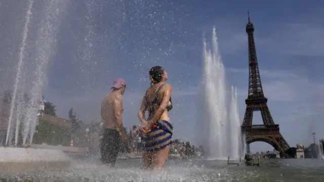 Two young people in swimming suits and bikini stand with their feet and legs partially submerged in a water fountain overlooking Paris' Trocadero, the Eiffel Tower in the background