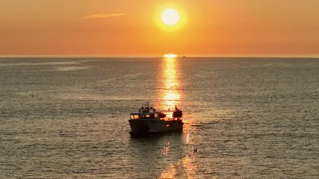 A small boat floats in the middle of a vast body of water as the sun rises on the horizon