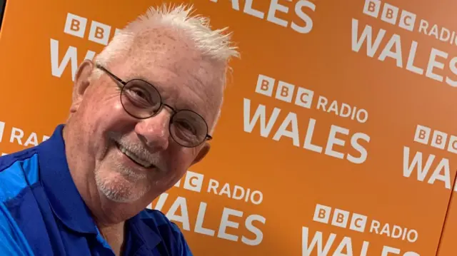A man smiles while facing the camera. He wears a bright blue shirt and is standing in front of an orange background screen, which has the logo for BBC Radio Wales in a repeating pattern