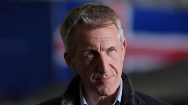 Dan Jarvis wears a jumper and a blue shirt and stands in front of a blurry Union Jack flag