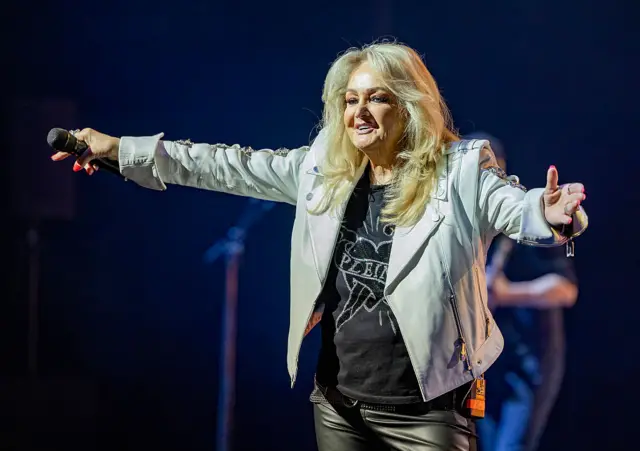Bonnie Tyler performing on stage with a smile on her face and arms open wide. She holds a microphone in her right hand.