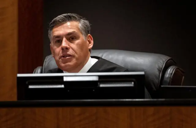 A judge in black robes and a white shirt sits at the bench in court