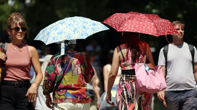 People walking and holding a blue and white and a red umbrella protecting themselves from the sun
