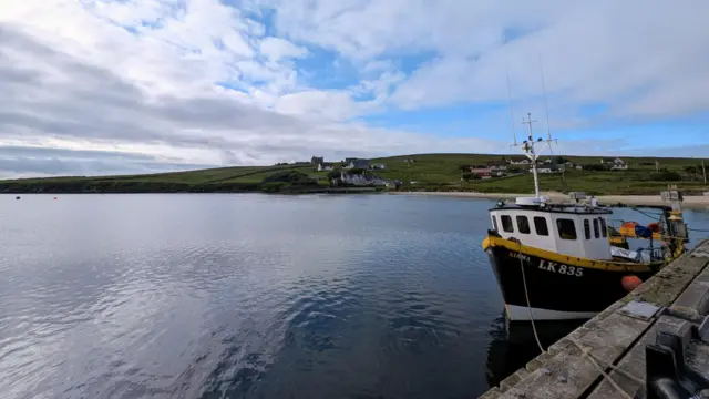 A boat is moored in the water next to a wooden jetty under a cloudy sky. More of the coast can be seen in the background, with a beach and large swathes of green fields