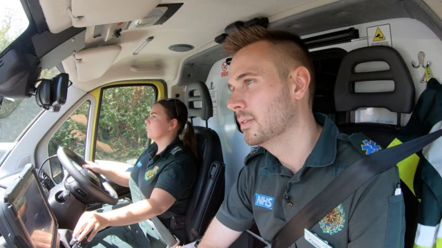 A view inside the drivers cab on an ambulance. The photo appears to be taken from the corner of the cab, closest to the windscreen, and shows one male paramedic sat in the passenger seat while a female paramedic sits in the driving seat next to him