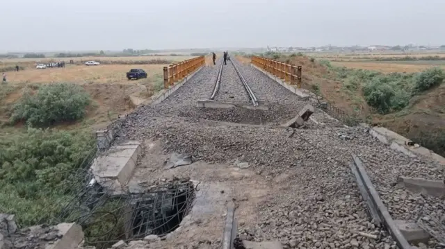A damaged railway bridge, the tracks have been split by a large crater, a large section of railings are destroyed and there is a hole in the side of it
